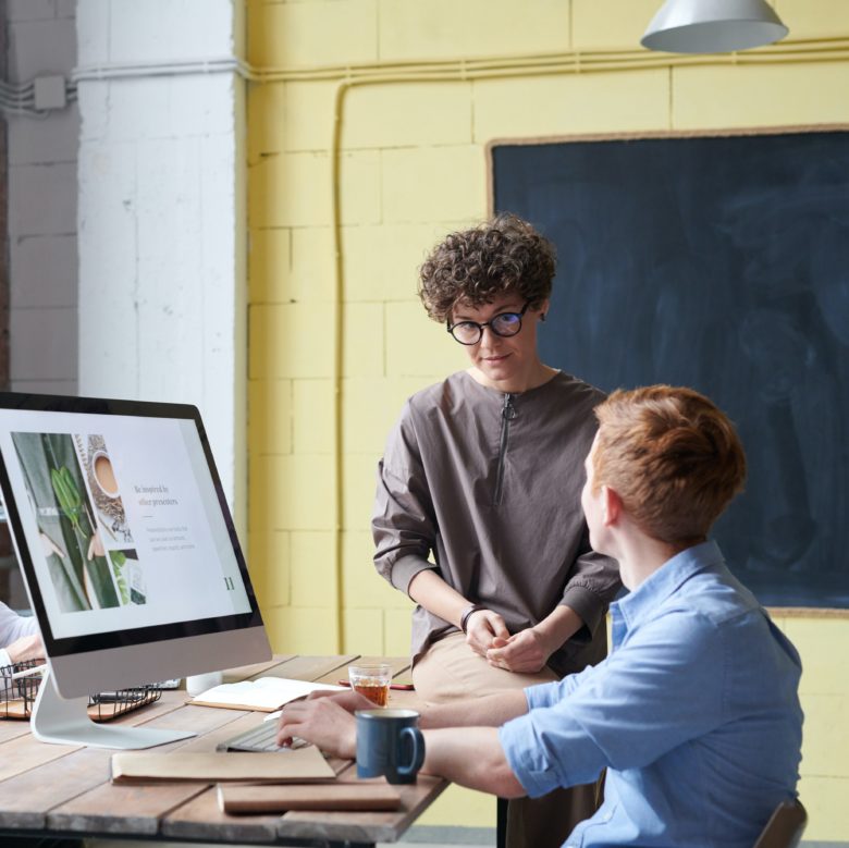 Jeune homme assis devant son ordinateur et discutant avec une jeune femme assise sur le bureau à côté de lui durant une séance de coaching professionnel.